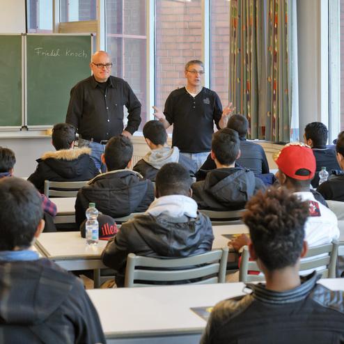 Zwei M&auml;nner stehen vorne vor einer Gruppe von Jugendlichen in einem Klassenraum mit gr&uuml;ner Tafel und Fenstern.