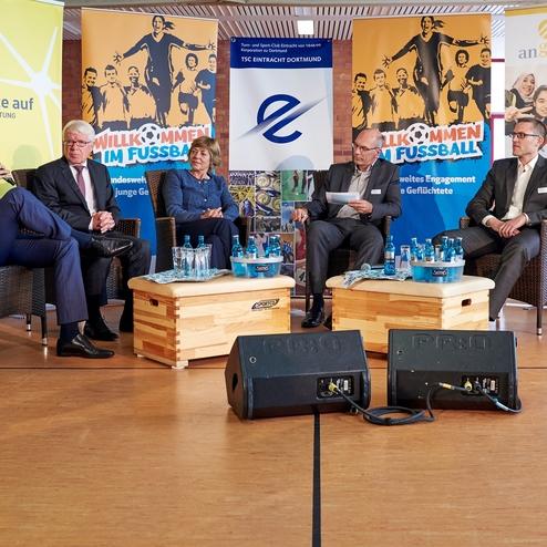 Podiumsdiskussion mit f&uuml;nf Personen auf einem Tisch, umgeben von Bannern &uuml;ber Fu&szlig;ball und Integration, in einem modernen Raum.