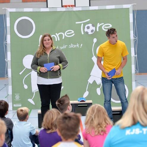 Zwei Personen stehen vor einer Sportwand und präsentieren aktiv vor einer Gruppe von Kindern in einem Turnsaal.