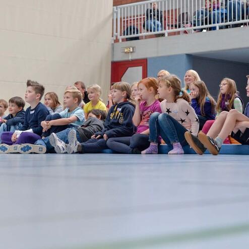 Eine Gruppe von Kindern sitzt auf dem Boden eines Sporthallenbodens und schaut aufmerksam in eine Richtung.