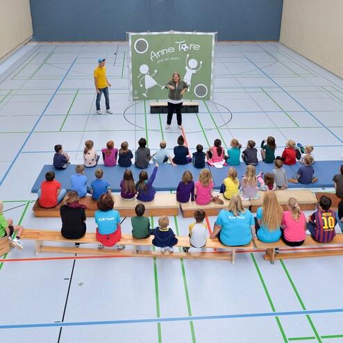 Gruppierung von Kindern auf Holzbänken in einer Sporthalle, während eine Person auf der Bühne spricht.