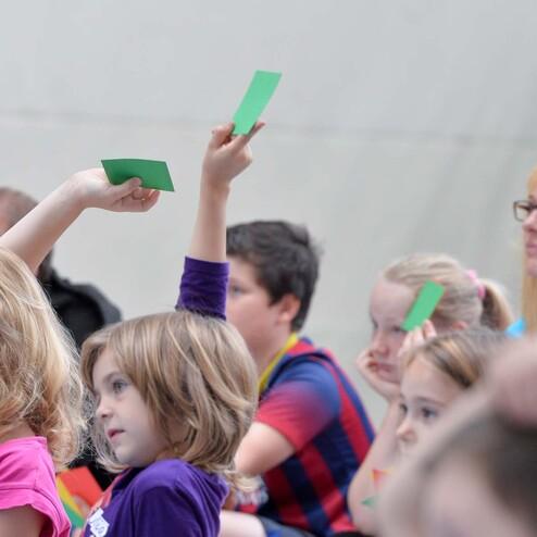 Gruppe von Kindern hebt grüne Karten in die Höhe, während ein Erwachsener im Hintergrund zuschaut.
