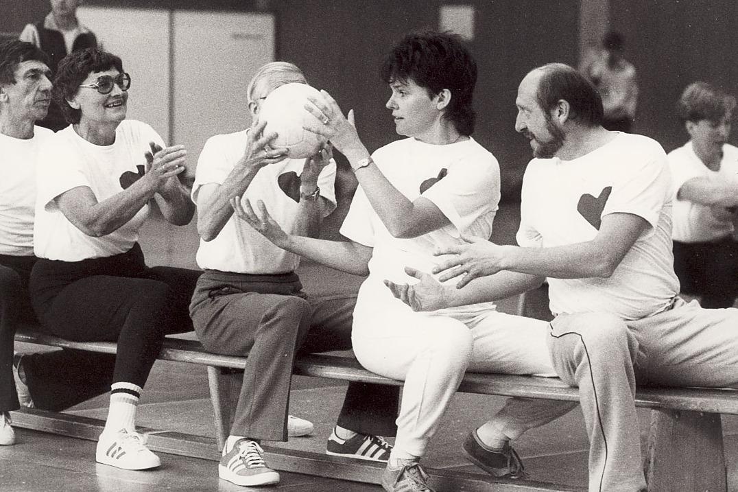 Gruppe von f&uuml;nf Menschen sitzt auf einer Bank, h&auml;lt einen Ball und tr&auml;gt T-Shirts mit Herzmotiven in einer Sporthalle.