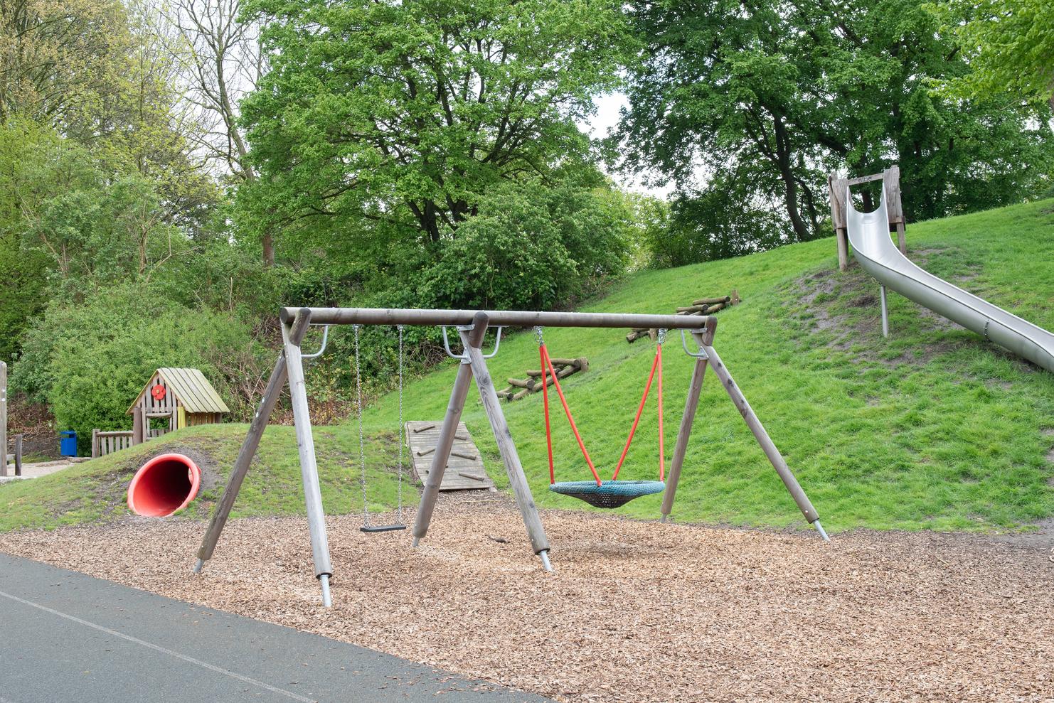 Spielplatz mit einer Schaukel, einer Rutsche und einem Spielhaus, umgeben von Rasen und B&auml;umen.