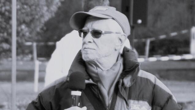 Älterer Mann mit Brille und Baseballmütze hält ein Mikrofon, blickt nachdenklich in die Ferne. Schwarz-Weiß-Foto.