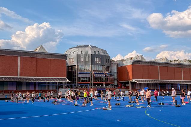 Menschen trainieren auf einem blauen Sportplatz vor einem modernen Geb&auml;udekomplex an einem sonnigen Tag mit Wolken.