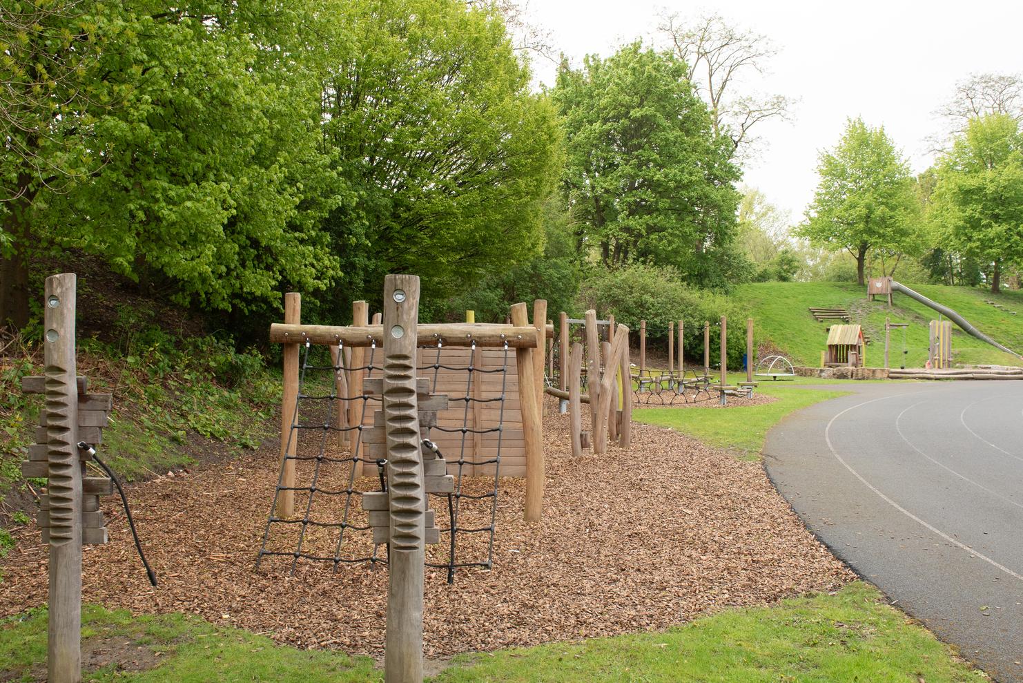 Spielplatzanlage mit Kletterger&uuml;st aus Holzelementen und angrenzender Laufbahn, umgeben von B&auml;umen und Grasfl&auml;chen.