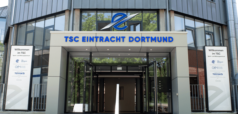 Eingang des TSC Eintracht Dortmund mit modernen Fenstern und Treppen, beschriftet mit "Willkommen im TSC".