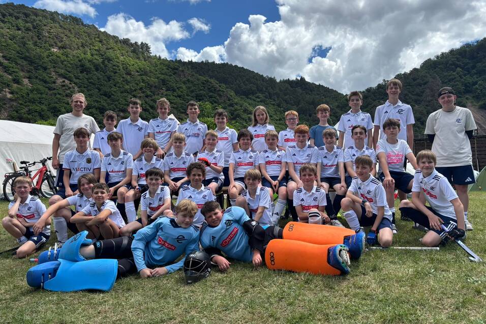 Gruppenfoto von 36 Jungen in Sportbekleidung mit Hockeyschl&auml;gern, auf einem Rasenfeld vor einer bergigen Landschaft.