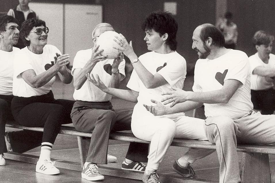 Gruppe von f&uuml;nf Menschen sitzt auf einer Bank, h&auml;lt einen Ball und tr&auml;gt T-Shirts mit Herzmotiven in einer Sporthalle.