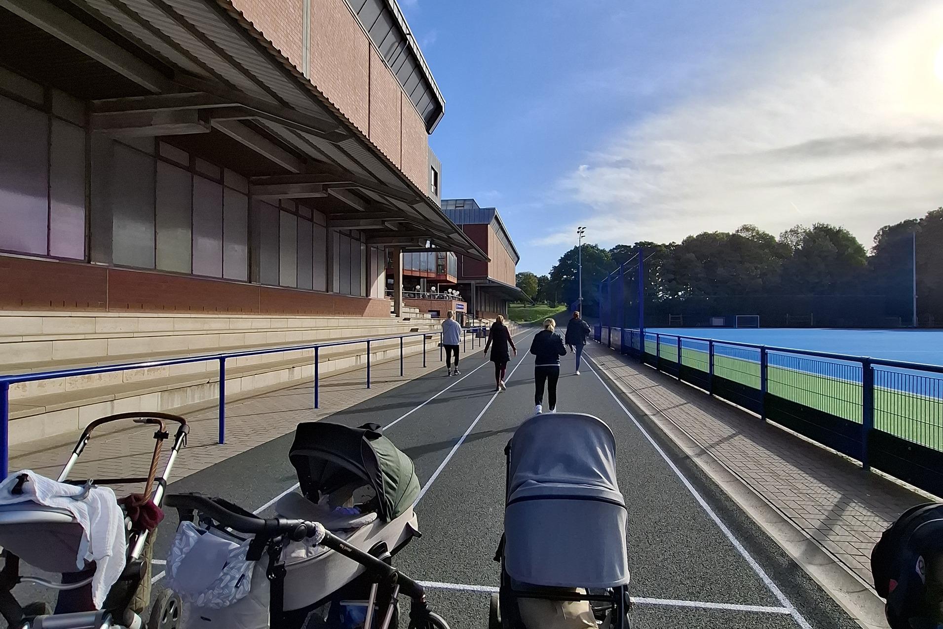 Weg mit mehreren Kinderwagen, während Personen entlang eines Sportgeländes spazieren. Helle Tagesstimmung, blaue Himmel.