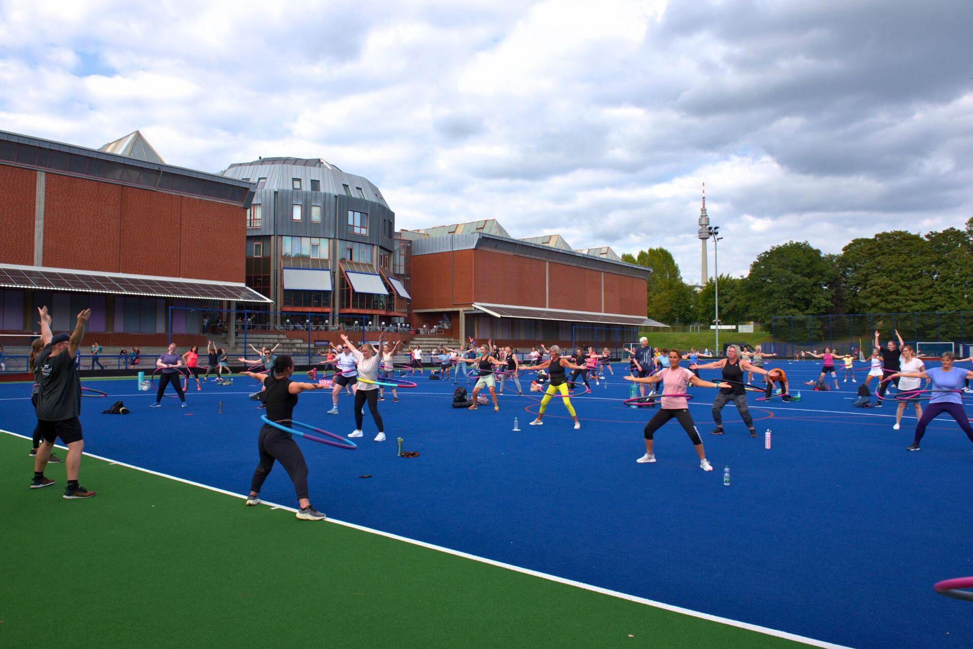 Gruppentraining auf einem blauen Sportplatz, Teilnehmer:innen nutzen Hula-Hoops und Fitnessgeräte unter bewölktem Himmel.