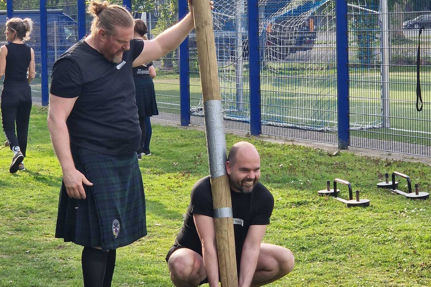 Zwei M&auml;nner &uuml;ben im Freien an einem Holzbalken; einer h&auml;lt den Balken, der andere beugt sich zum Anheben.