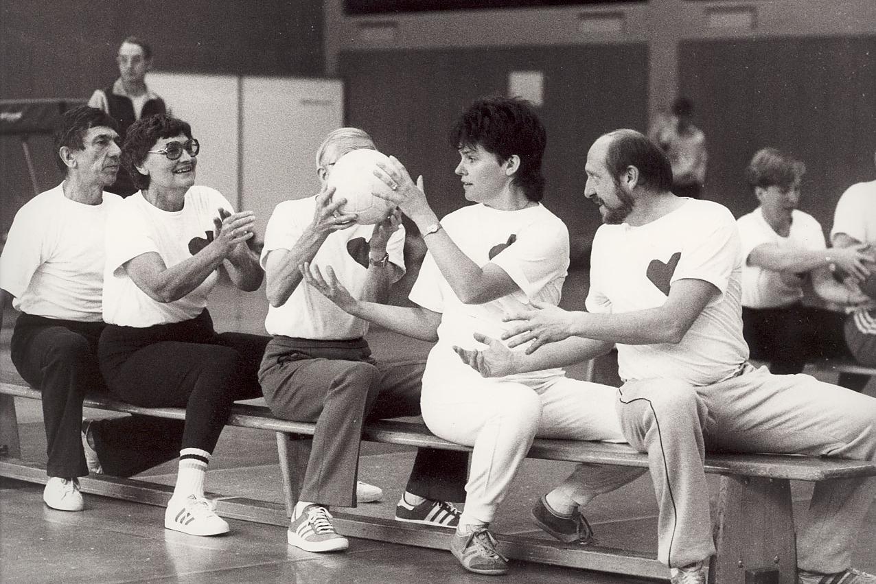 Gruppe von f&uuml;nf Menschen sitzt auf einer Bank, h&auml;lt einen Ball und tr&auml;gt T-Shirts mit Herzmotiven in einer Sporthalle.