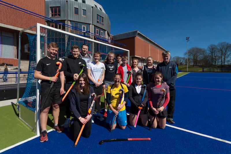 Gruppenshot von 16 Hockeyspielern mit Schlägern auf einem blauen Platz vor einem Sportgebäude an einem sonnigen Tag.