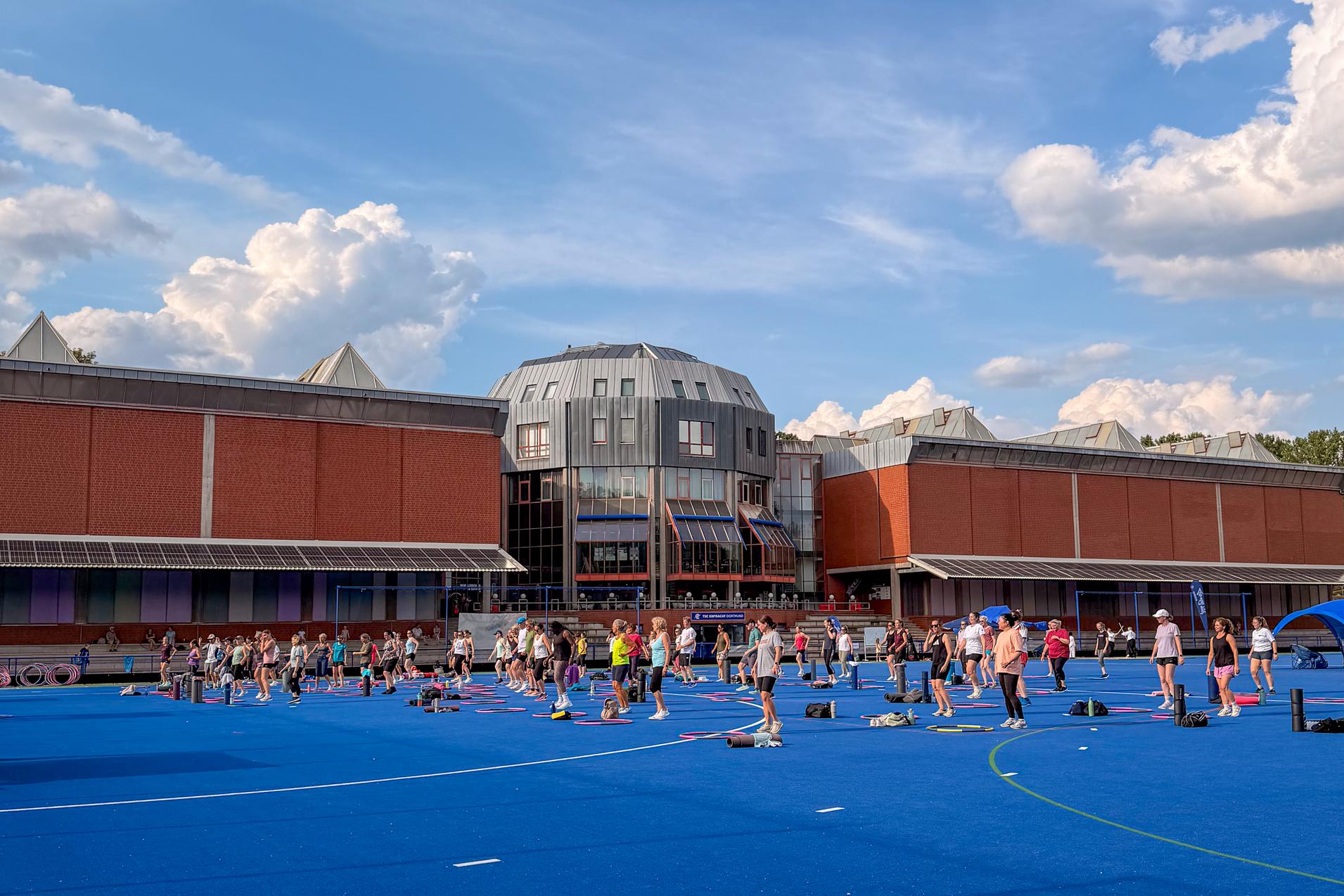 Menschen trainieren auf einem blauen Sportplatz vor einem modernen Gebäudekomplex an einem sonnigen Tag mit Wolken.