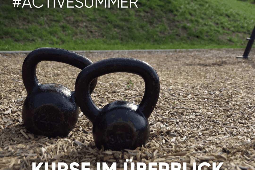 Zwei schwarze Kettlebells liegen auf einem Holzschnitzelboden im Freien. Im Hintergrund ist eine grüne Wiese sichtbar.