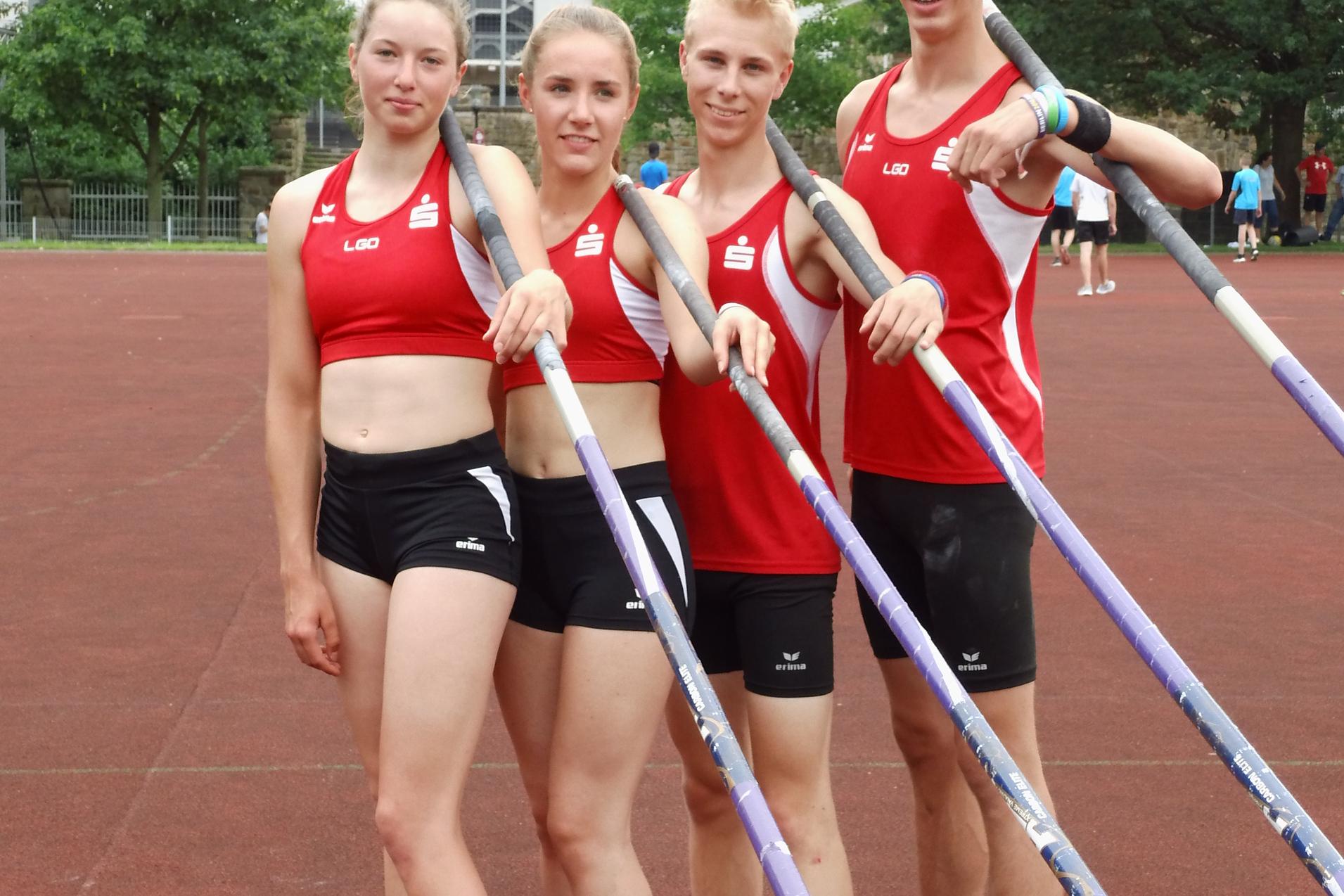 Vier Sportler in roten Trikots stehen auf einer Laufbahn und halten Stabhochsprungstangen, im Hintergrund eine Sportstätte.