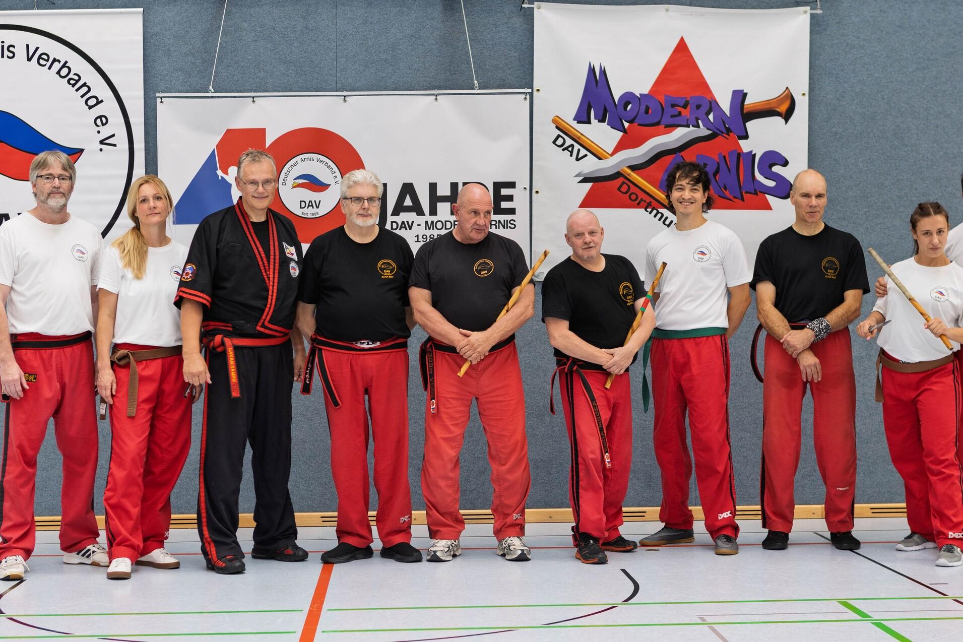 Gruppe von neun Personen in roten Hosen und T-Shirts, posierend in einem Sportstudio vor Bannern des Deutschen Arnis Verbands.
