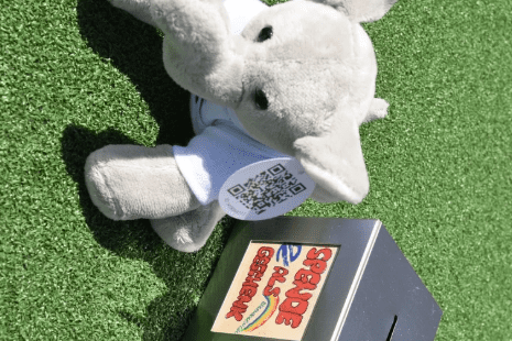 Pl&uuml;sch-Elefant mit QR-Code auf einem gr&uuml;nen Untergrund neben einer silbernen Spendenbox.