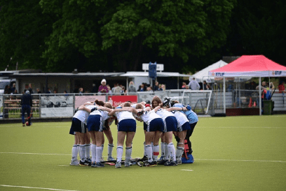 Mannschaftsmitglieder in blau-weißen Hockeytrikots bilden einen engen Kreis auf dem Hockeyfeld.