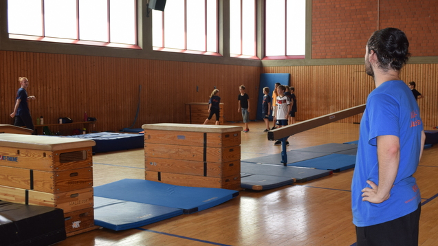 Zwei Trainer beobachten eine Trainingsgruppe in einer Sporthalle mit mehreren &Uuml;bungsger&auml;ten und Matten.