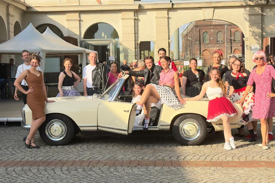Gruppe von Menschen in Retro-Kleidung posiert um ein wei&szlig;es Oldtimer-Cabrio auf einem Platz.