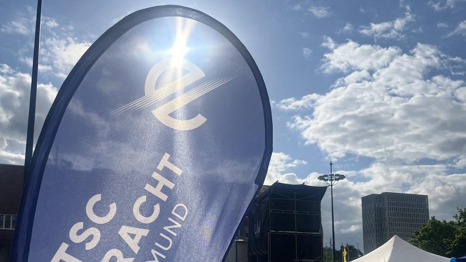 Fahne mit der Aufschrift „TSC Eintracht Dortmund“ im Vordergrund, während Menschen im Hintergrund an einem Event teilnehmen.