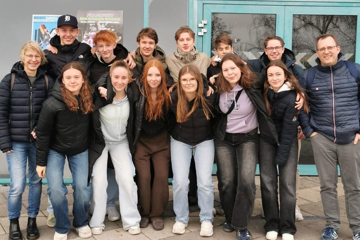 Gruppe von zwölf Personen steht lächelnd vor einer Glasfassade, während einige auf den Boden schauen.