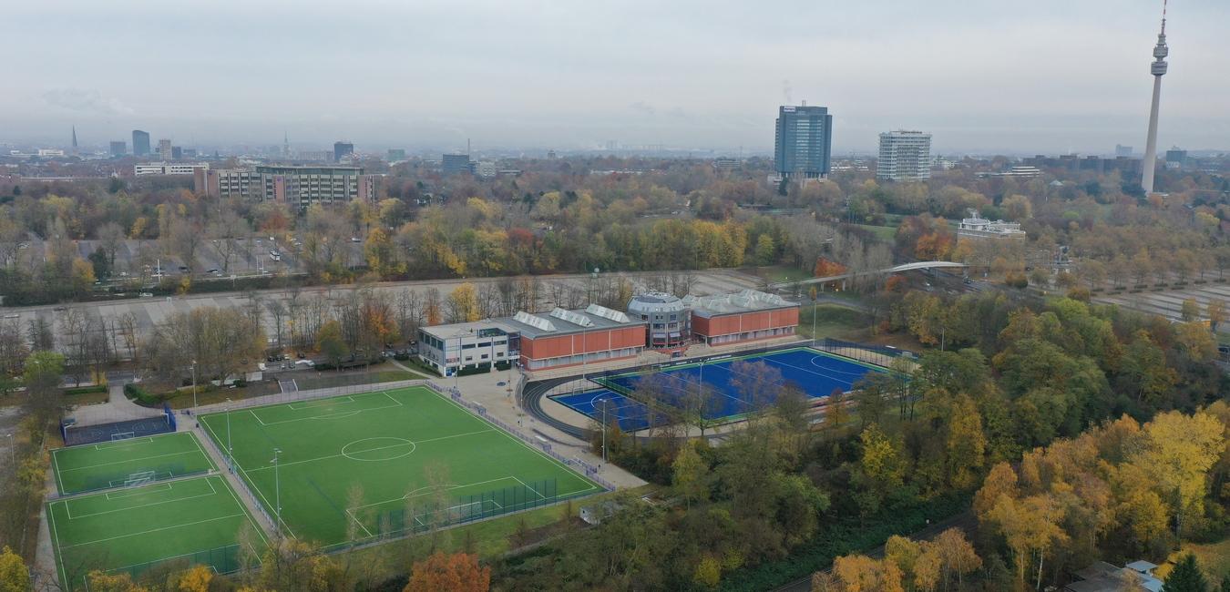 Luftaufnahme eines Sportkomplexes mit Fu&szlig;ballplatz, Schwimmbecken und umliegenden B&auml;umen im herbstlichen Stadtbild.