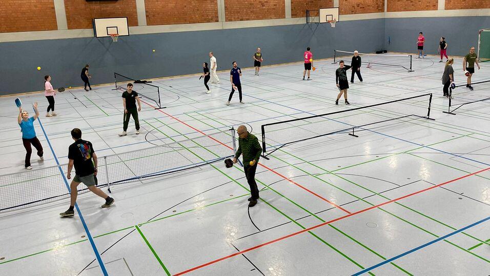 Vielzahl von Personen, die in einer Sporthalle Tennis spielen, umgeben von Netzen und bunten Linien auf dem Boden.