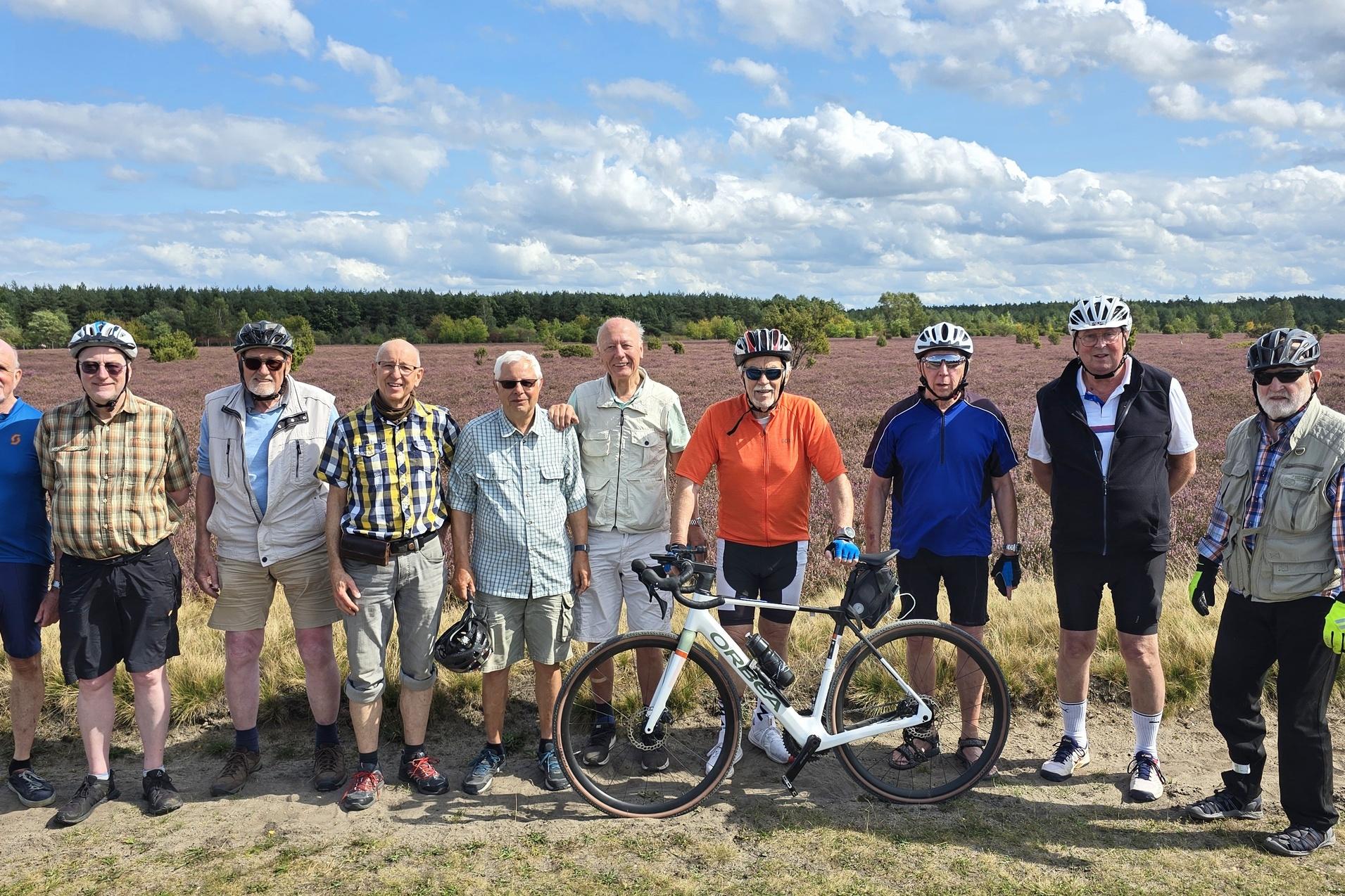 Gruppenshooting von zehn Radfahrern in Fahrradhelmen vor bl&uuml;hendem Heidefeld unter blauem Himmel.