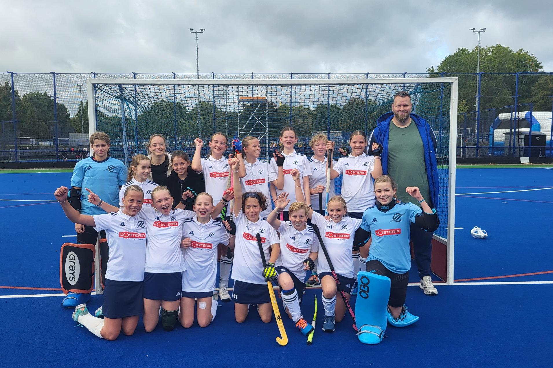 M&auml;dchenhockeyteam in wei&szlig;en Trikots posiert jubelnd vor einem Tor auf einem blauen Kunstrasenplatz.