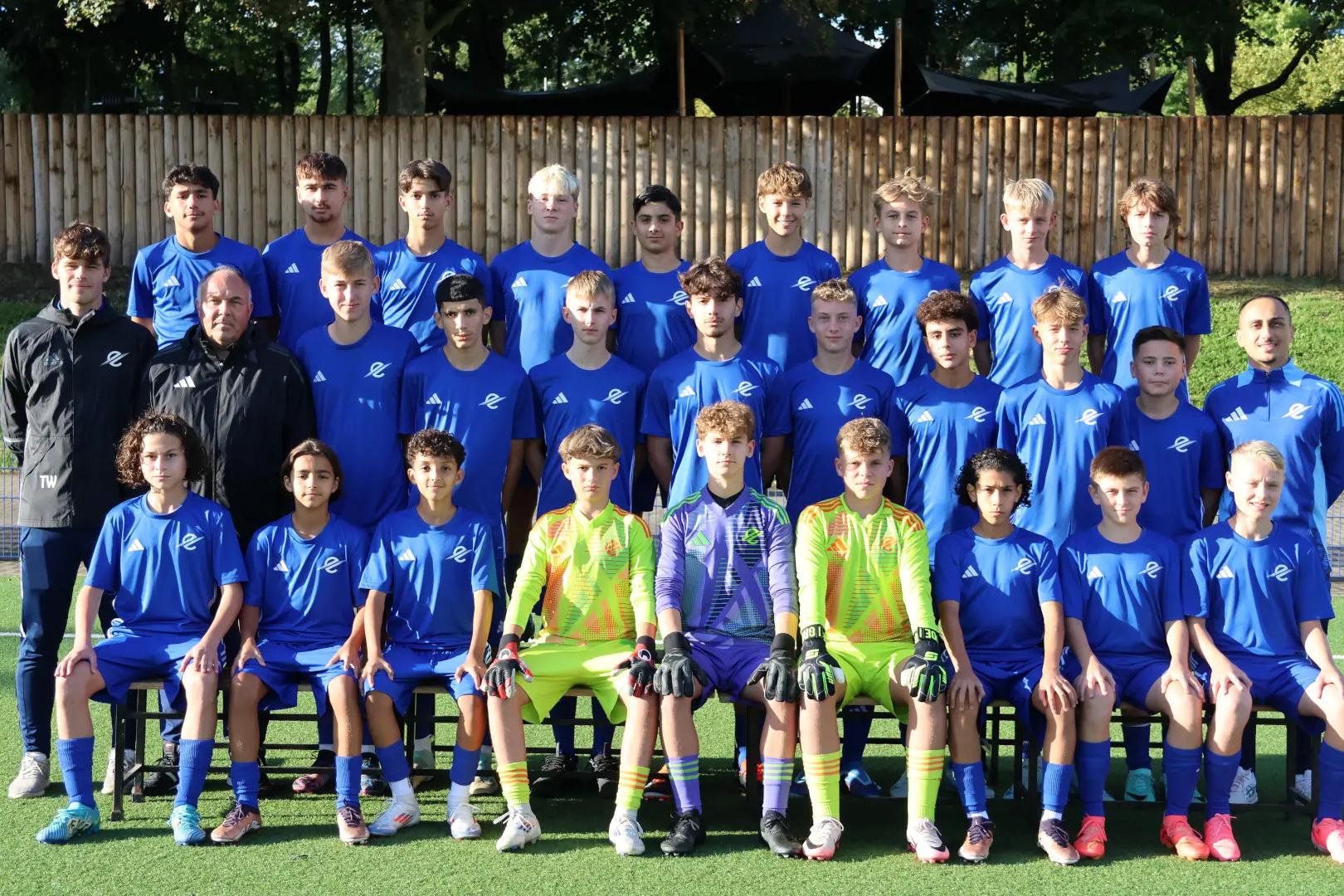 Teamfoto der C1 des TSC Eintracht Dortmund. 28 Personen in drei Reihen auf einem Kunstrasenplatz, alle in blauen Trikots, drei Torh&uuml;ter in neonfarbenen Outfits. Zwei Trainer au&szlig;en. Im Hintergrund: Holzzaun und B&auml;ume bei Tageslicht.