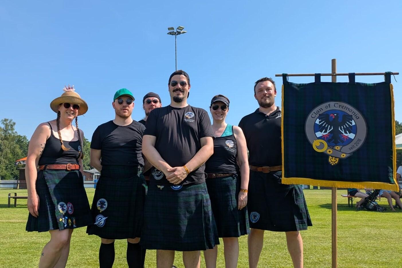 Sechs Personen in Kilts stehen auf einer Wiese neben einem Fahnenmast mit Banner des Clans. Sonnenlichter und blauer Himmel.