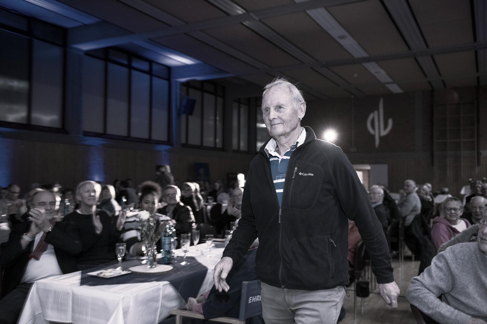 Ein &auml;lterer Mann geht l&auml;chelnd durch einen festlich dekorierten Saal, w&auml;hrend das Publikum applaudiert.
