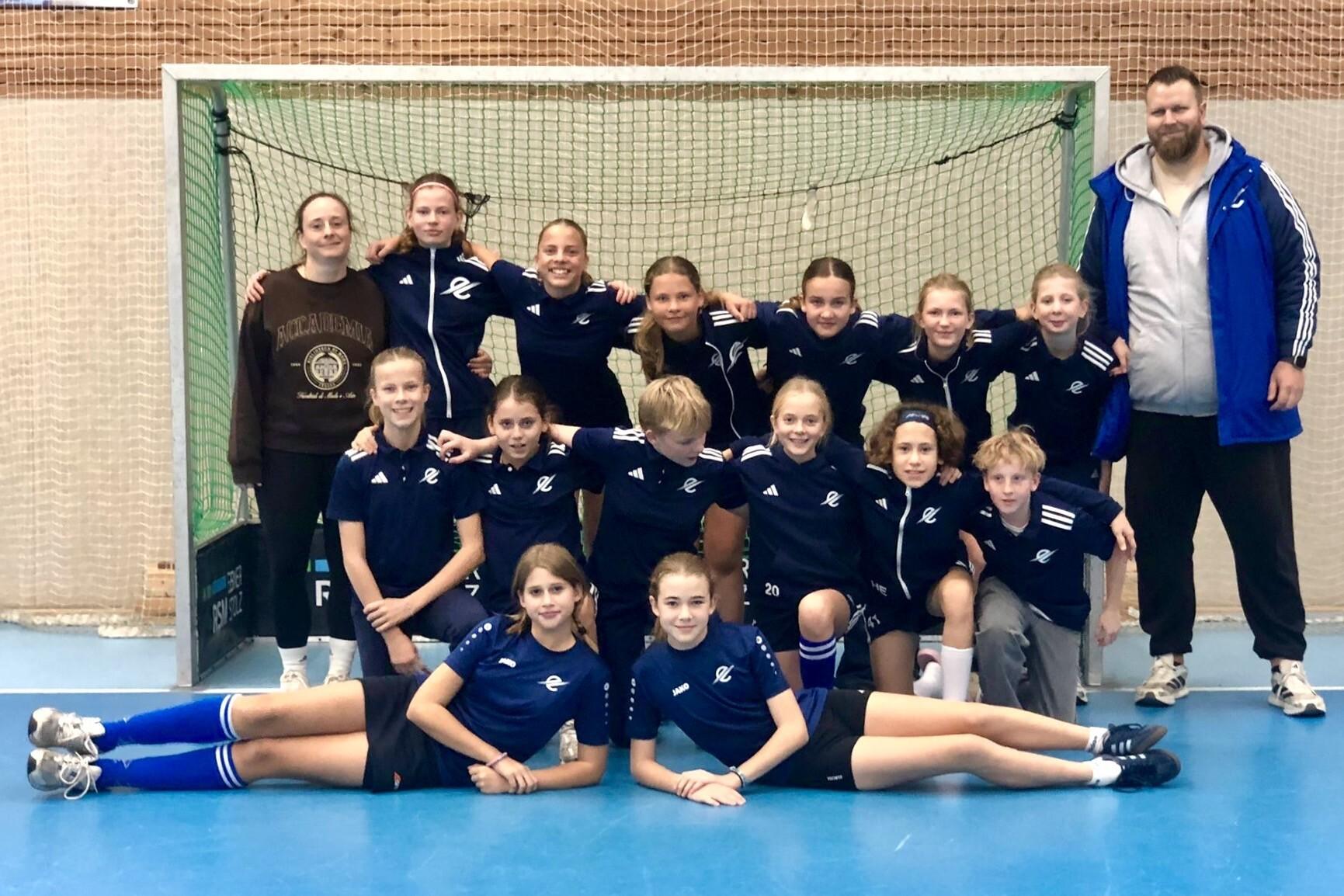 Mannschaftsfoto einer M&auml;dchen-Handballmannschaft mit Trainer vor einem Tor in einer Sporthalle.