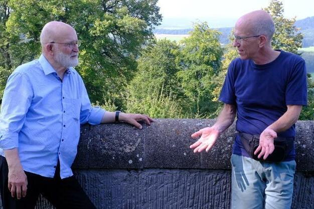 Zwei Männer stehen an einer Steinmauer und unterhalten sich vor einer grünen Landschaft mit Bäumen und Hügeln.