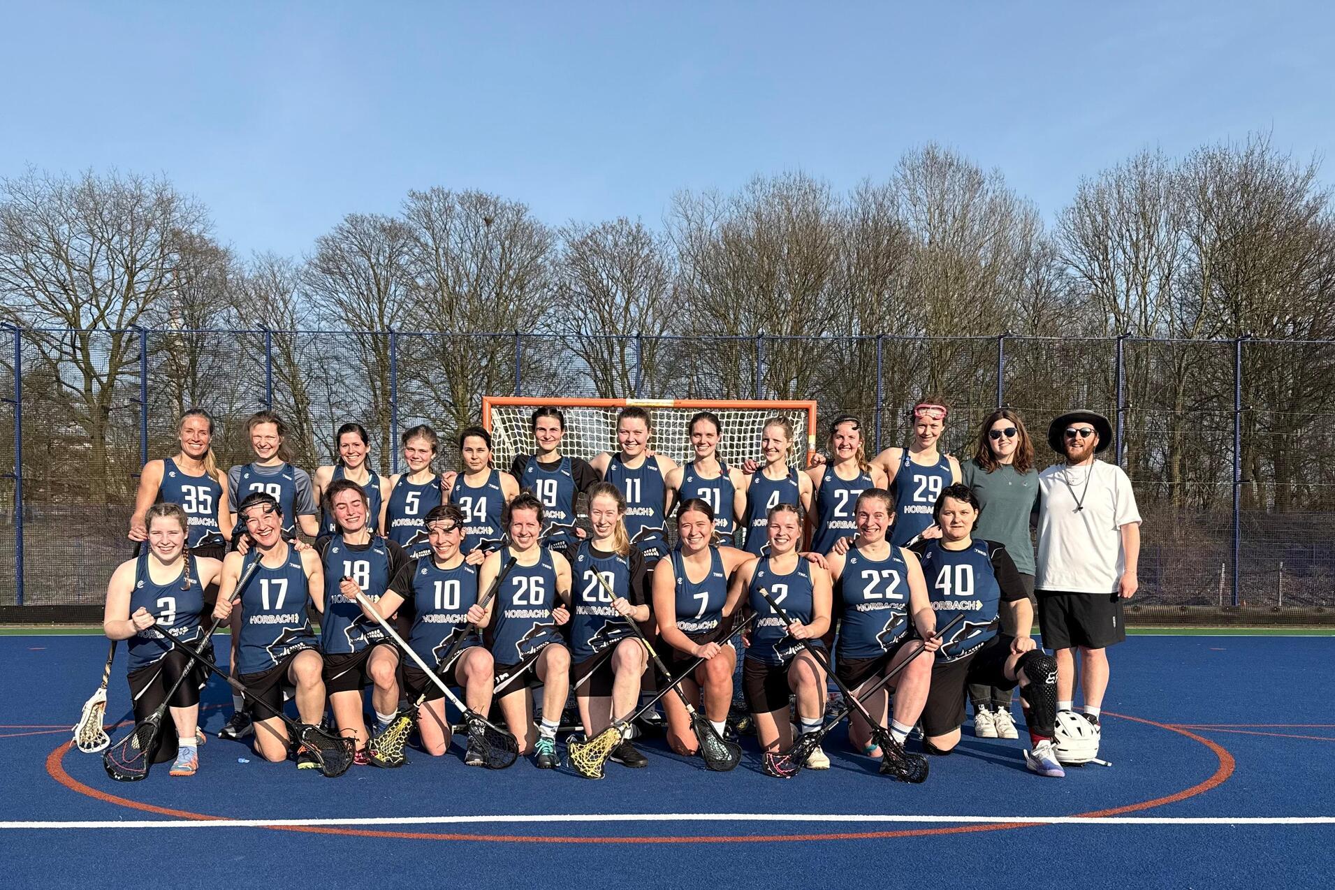Gruppierung von 22 Lacrosse-Spielerinnen in blauen Trikots auf einem Feld mit Tor im Hintergrund, bei sonnigem Wetter.