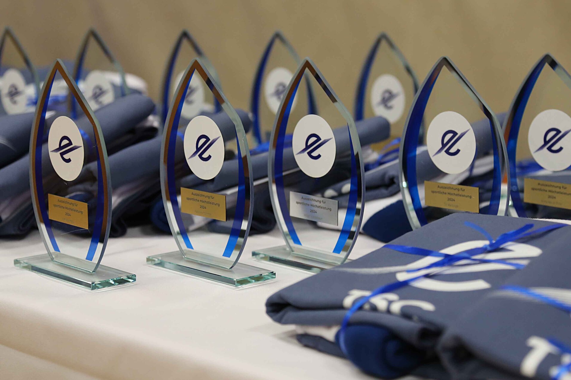 Troph&auml;en in Tropfenform mit blauen Akzenten stehen neben gestapelten T-Shirts auf einem Tisch.