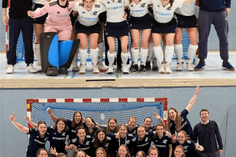 Zwei Gruppen von Frauenhockeyspielern posieren freudig vor Toren in einer Sporthalle.