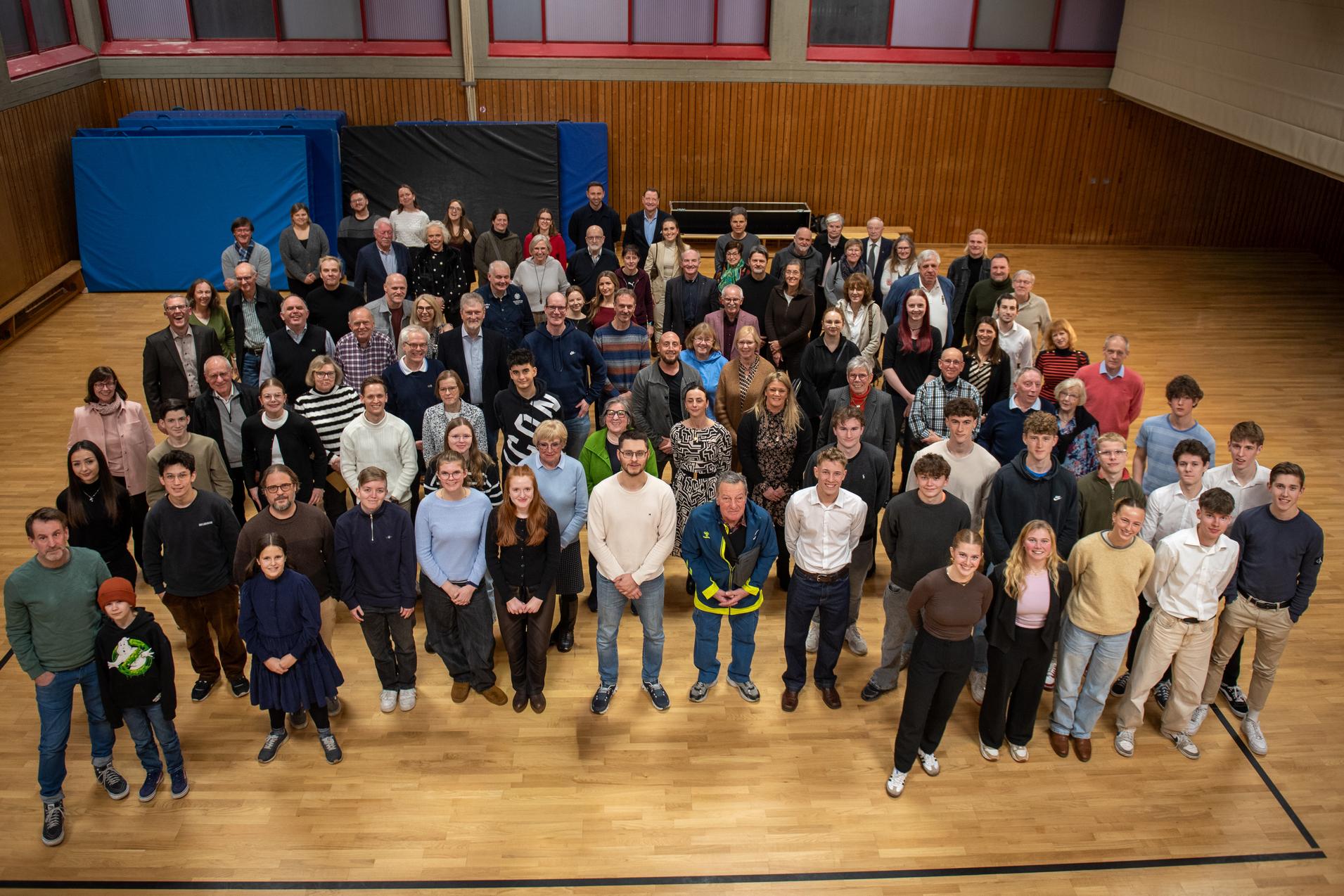 Gruppeneinheit von &uuml;ber 100 Personen in einem Sportraum, verteilt auf einer Holzfl&auml;che, einige halten B&auml;lle.