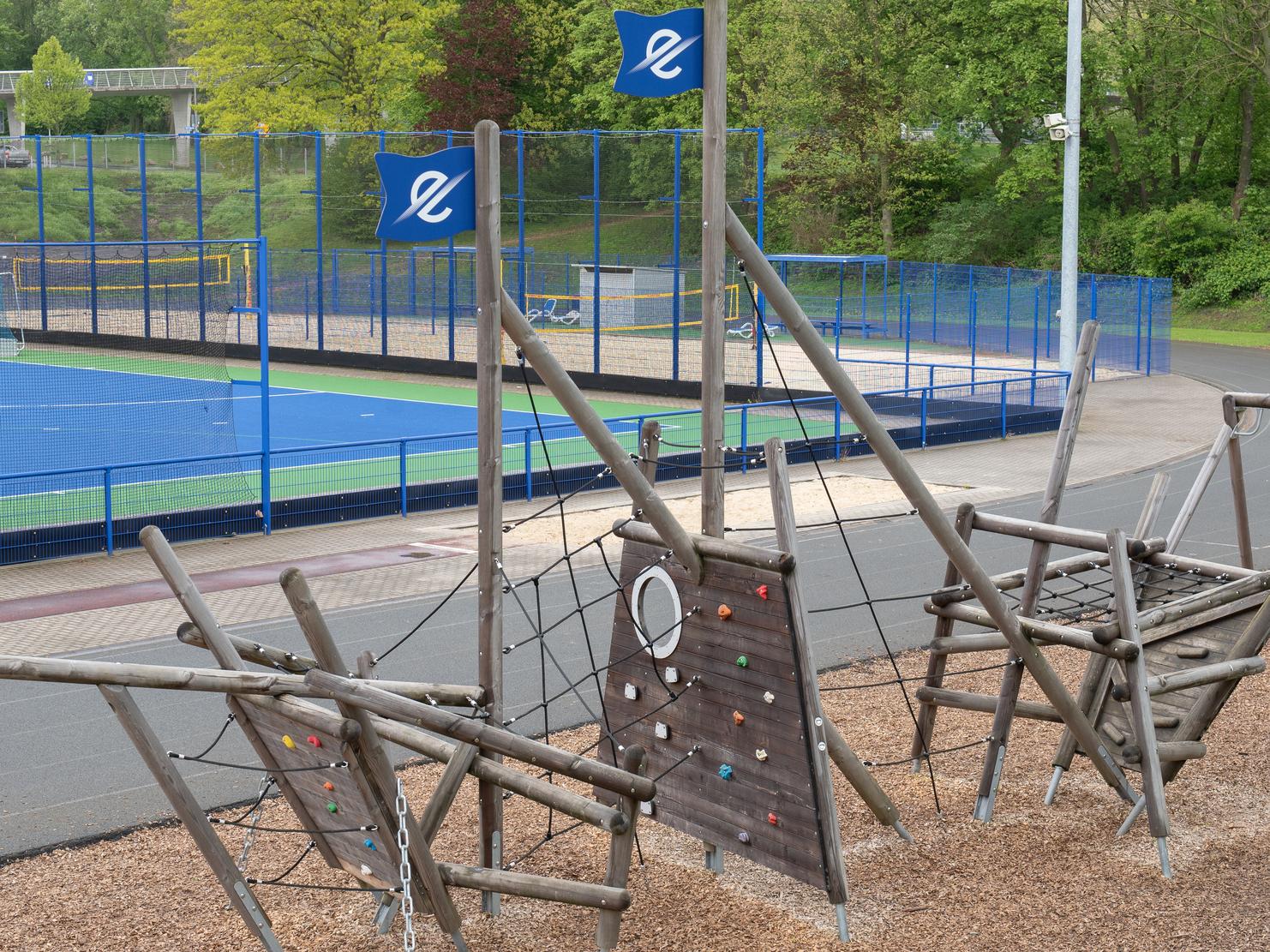 Spielplatz mit Kletterger&uuml;st aus Holz und Boulderwand, im Hintergrund ein Sportplatz und gr&uuml;ne Umgebung.