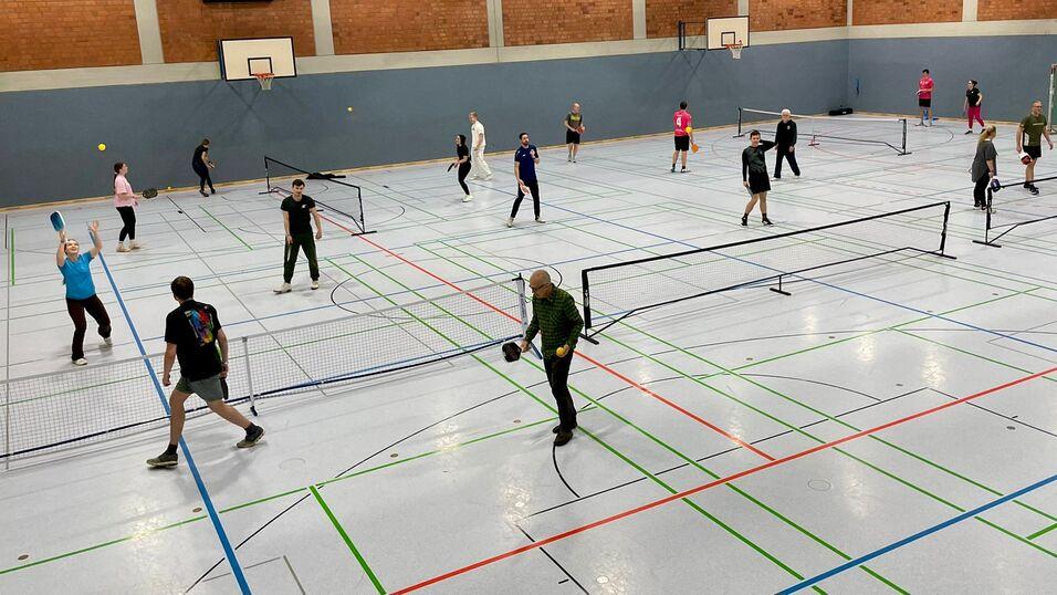 Vielzahl von Personen, die in einer Sporthalle Tennis spielen, umgeben von Netzen und bunten Linien auf dem Boden.