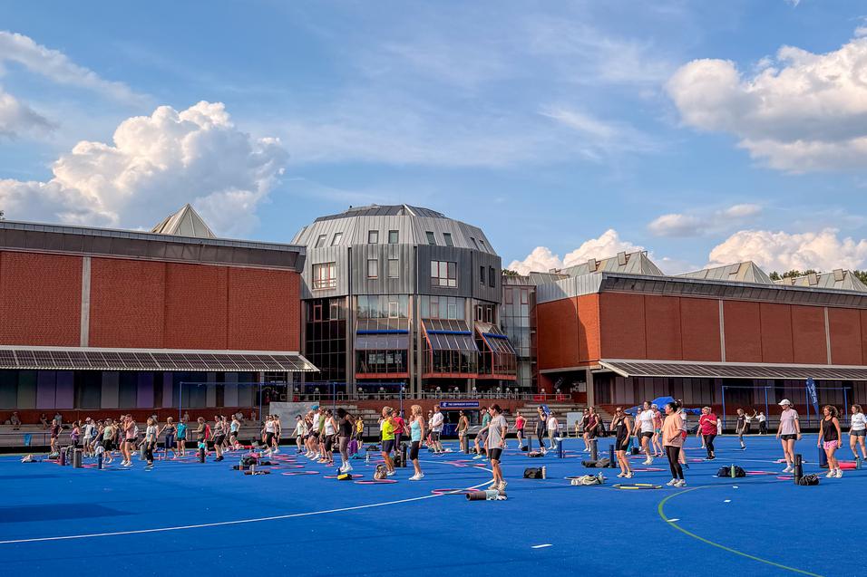 Menschen trainieren auf einem blauen Sportplatz vor einem modernen Geb&auml;udekomplex an einem sonnigen Tag mit Wolken.