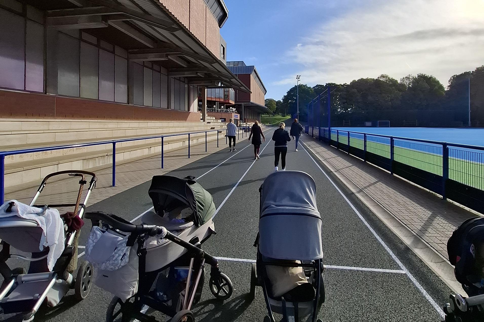 Weg mit mehreren Kinderwagen, w&auml;hrend Personen entlang eines Sportgel&auml;ndes spazieren. Helle Tagesstimmung, blaue Himmel.