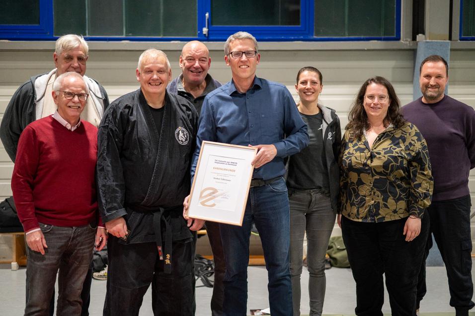 Gruppe von sieben Personen steht l&auml;chelnd zusammen, einer h&auml;lt eine Urkunde in der Hand, in einem Sportraum.