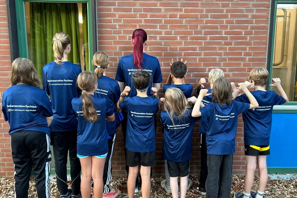Gruppe von Kindern und einem Erwachsenen in blauen T-Shirts, die von hinten in Richtung einer Wand schauen.