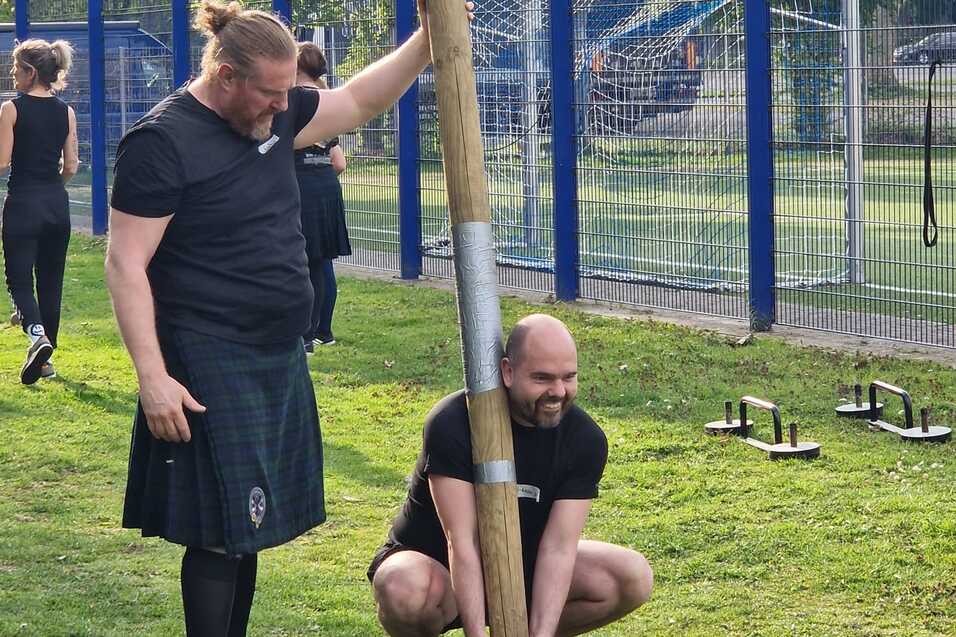Zwei M&auml;nner &uuml;ben im Freien an einem Holzbalken; einer h&auml;lt den Balken, der andere beugt sich zum Anheben.