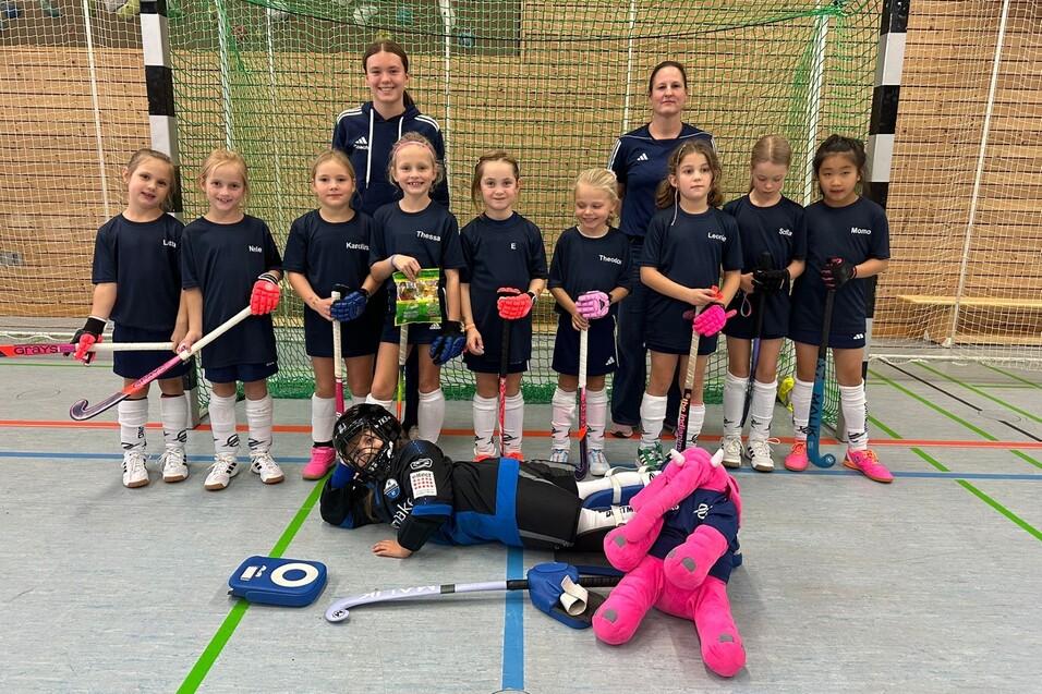 Gruppe von M&auml;dchen in Sportkleidung mit Schl&auml;gern und B&auml;llen, posed vor einem Hockeytor in einer Sporthalle.